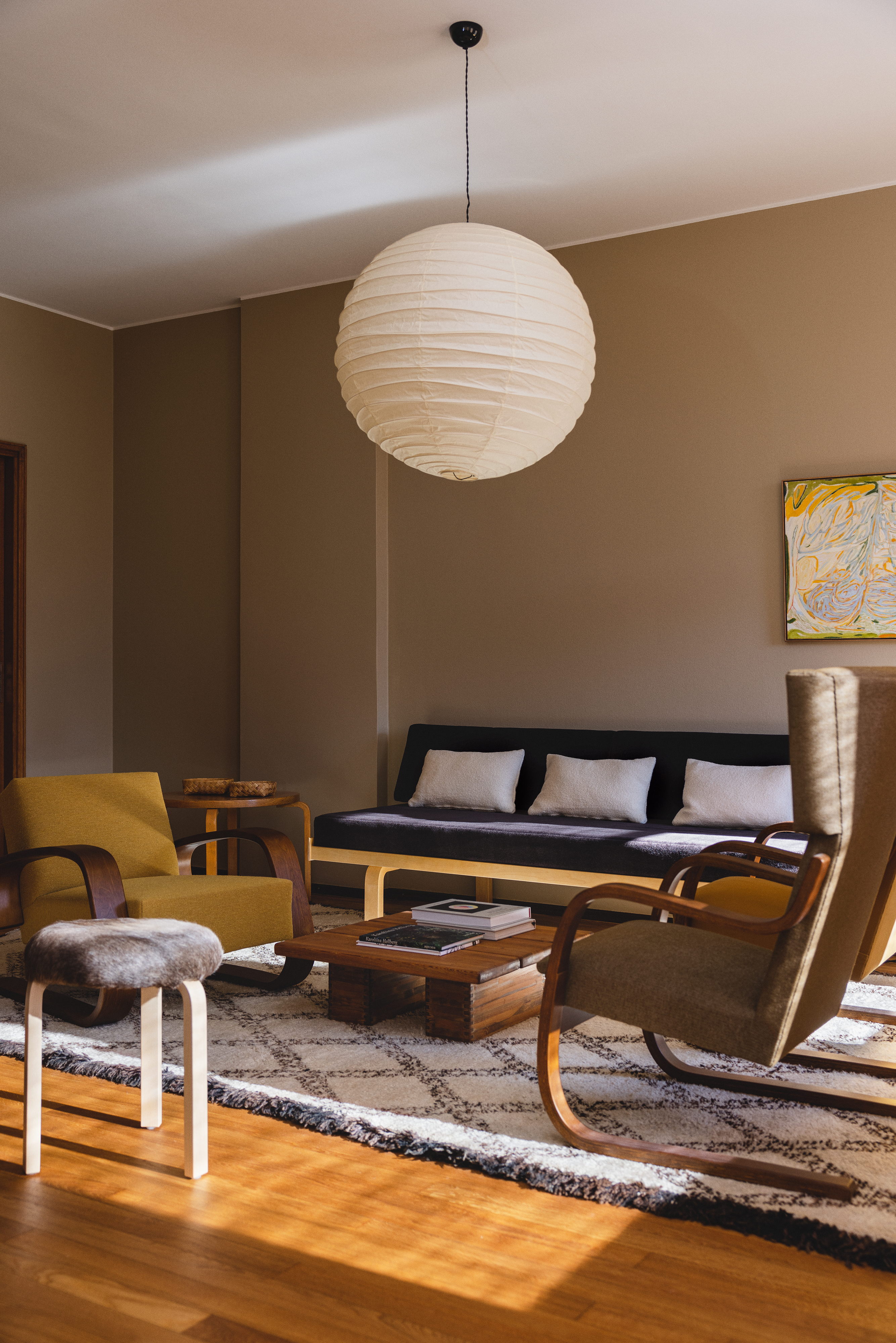 A modernist living room with sculptural wood and textile furniture in simple lines, a spherical paper lantern pending from the ceiling, wooden floors, checkered black and white rugs, and colorful art is brought to life by natural light.
