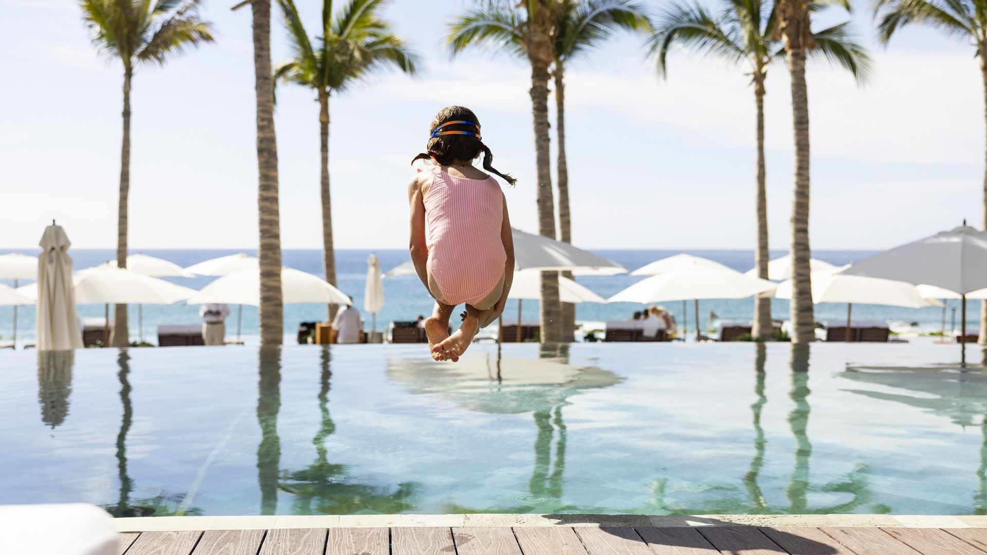 A young girl jumps into a pool, a beach and palm trees in the background.