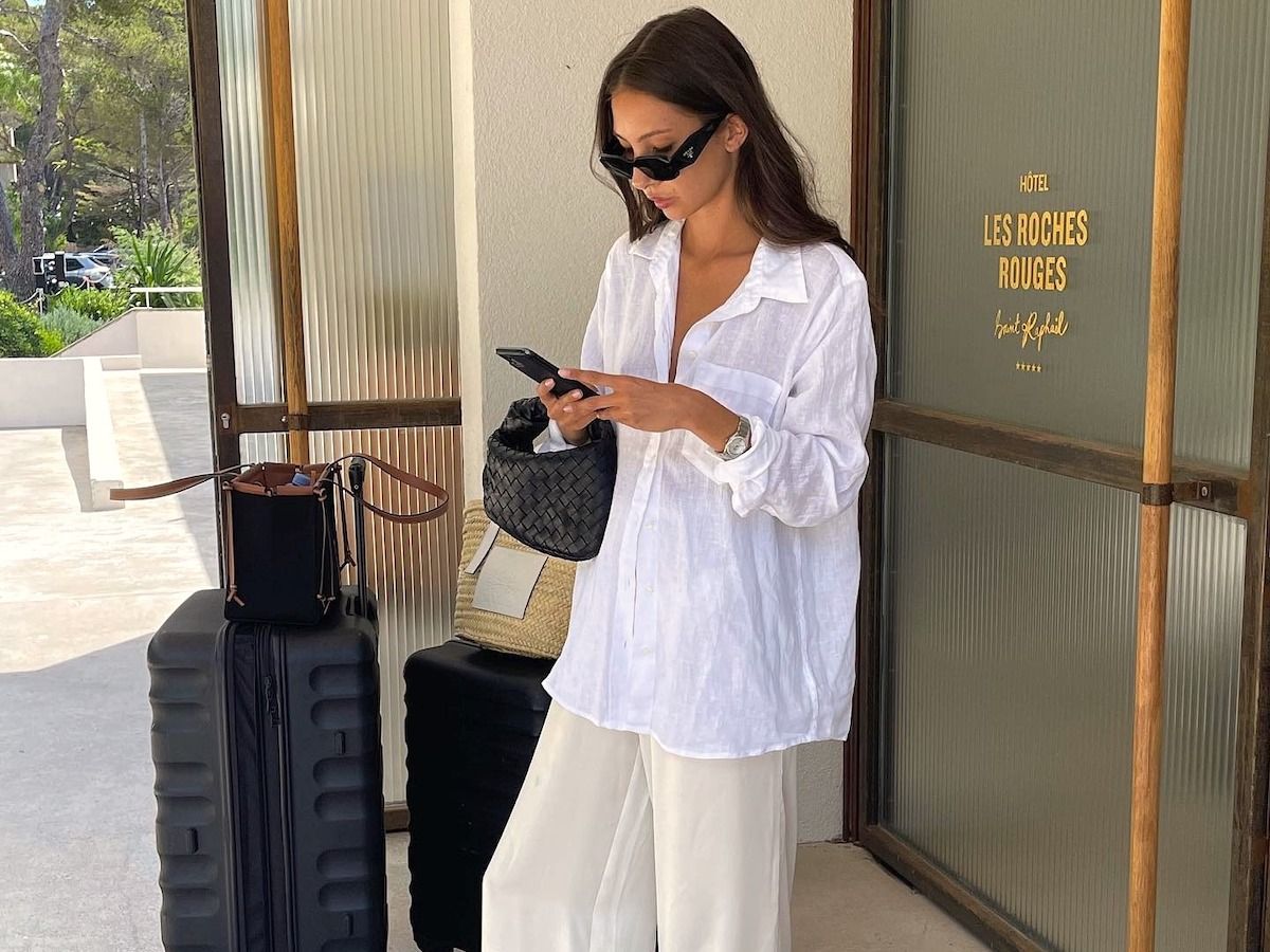 Style influencer Felicia Akerstrom poses with her suitcases while looking at her iPhone in front of the Les Roches Rouges hotel in France wearing black sunglasses, an oversize white linen button-down shirt, relaxed ivory silk pants, and a small black woven Bottega Veneta bag.