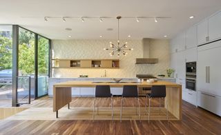 An open plan kitchen featuring a wooden design island, with 3 high chairs; Laminate flooring; Grey kitchen cabinets, silver appliances and a chandelier hanging from the ceiling