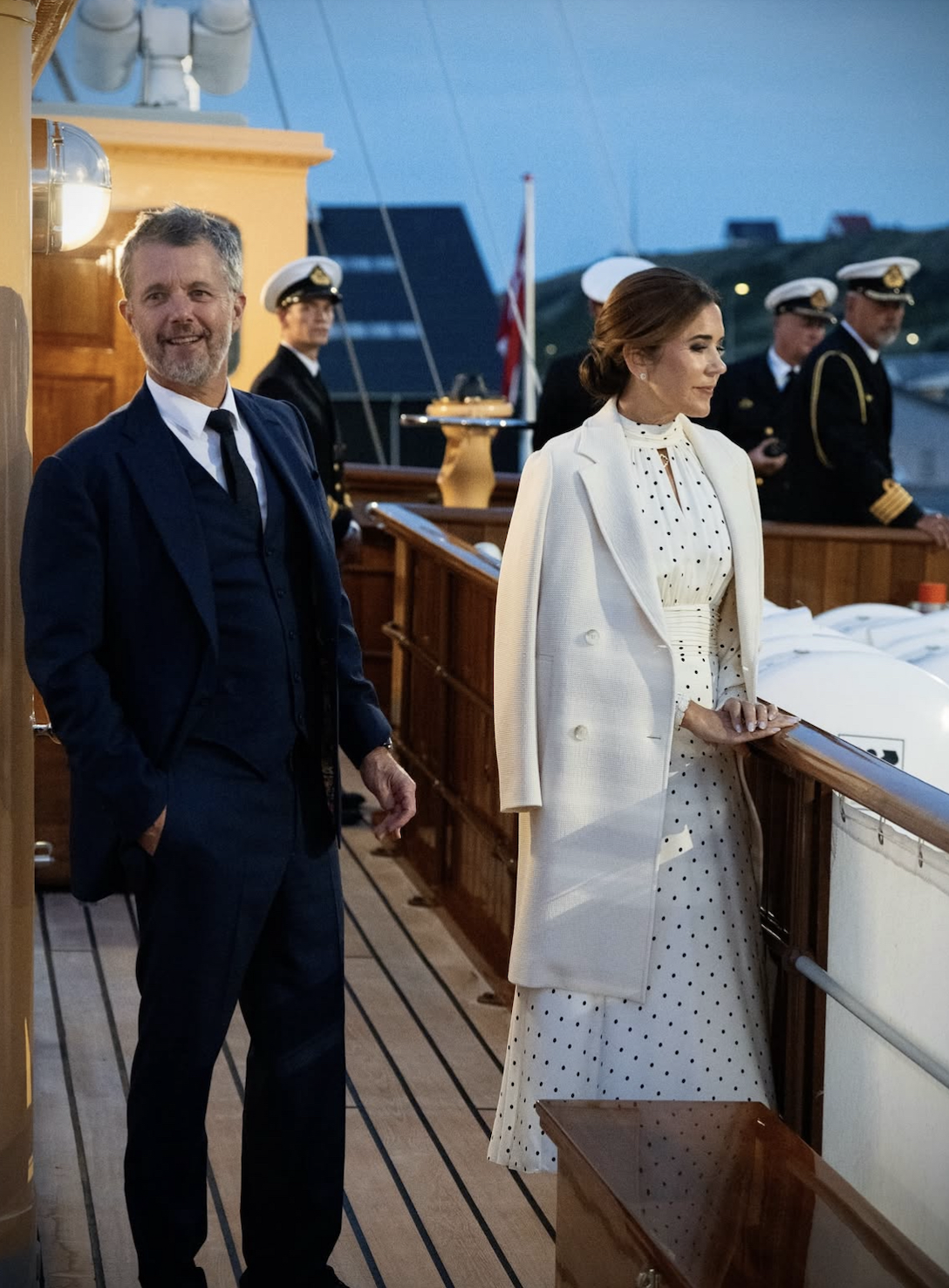 Queen Mary wearing a white polka dot dress and white coat and King Frederik wearing a suit aboard the royal yacht