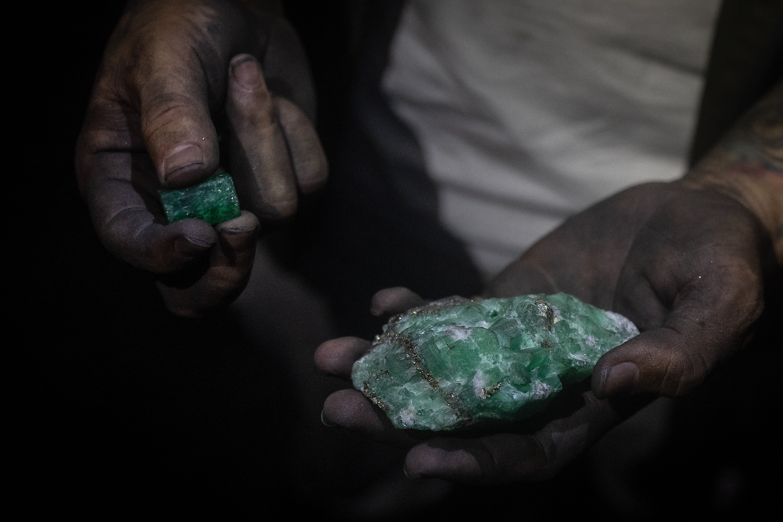 A close-up photograph of a miner's hands holding an uncut emerald