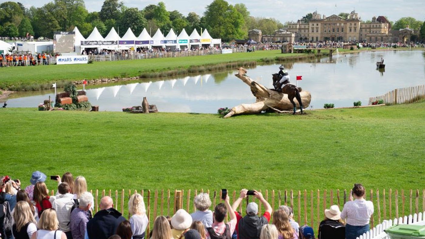 Image shows a horse jumping over a log into the lake at Badminton Horse Trials, with crowds in the foreground and a tented hospitality village in the distance