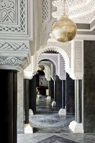 A hotel corridor with arched doorways, white pillars, dark grey walls and light grey patterned floor tiles.