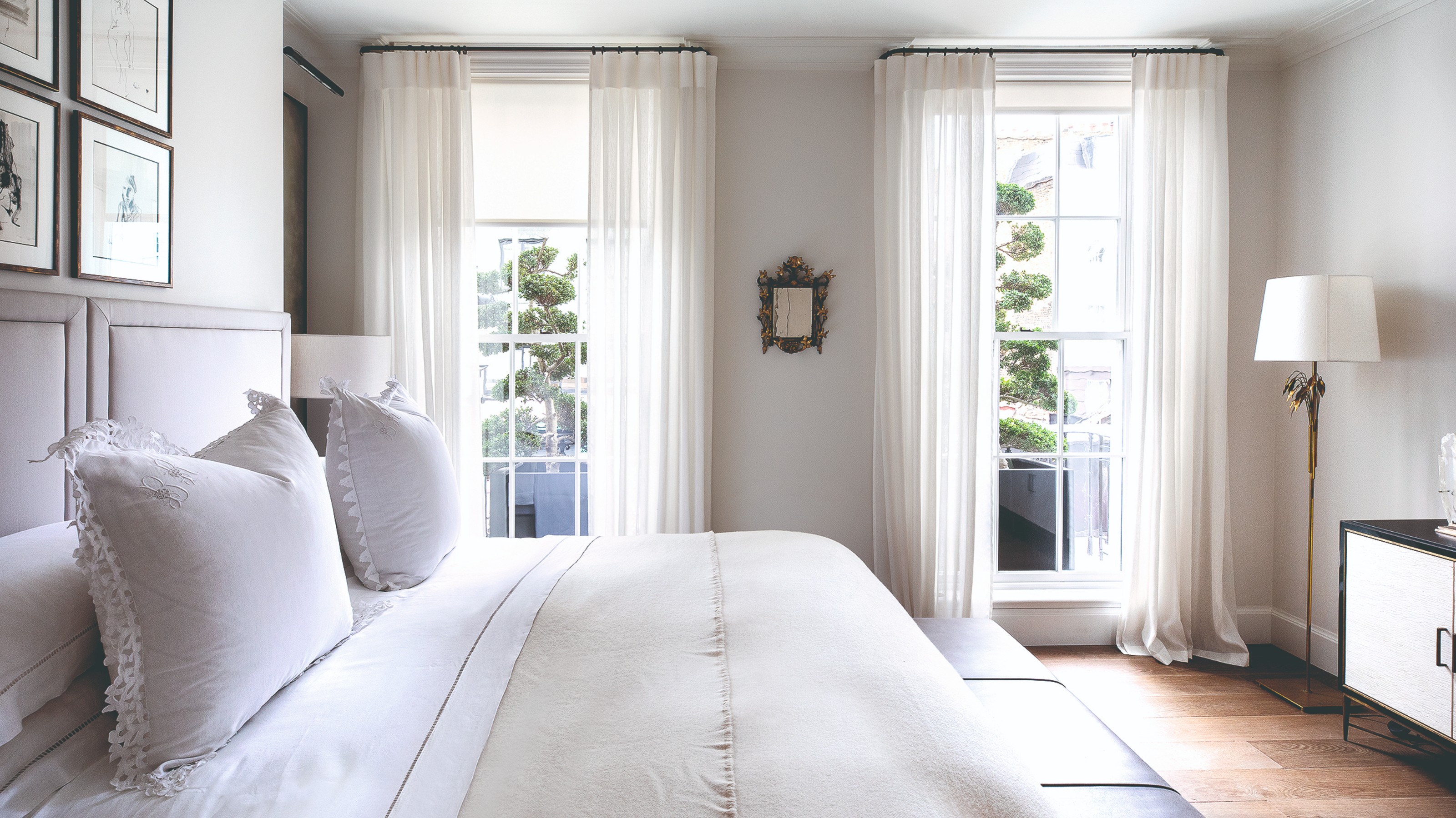 A white bedroom with two large windows dressed with white voile curtains and tonal roller blinds
