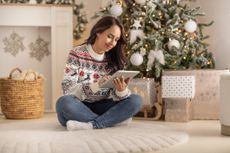 a woman sitting in front of a Christmas tree and shopping for Christmas presents on a tablet.