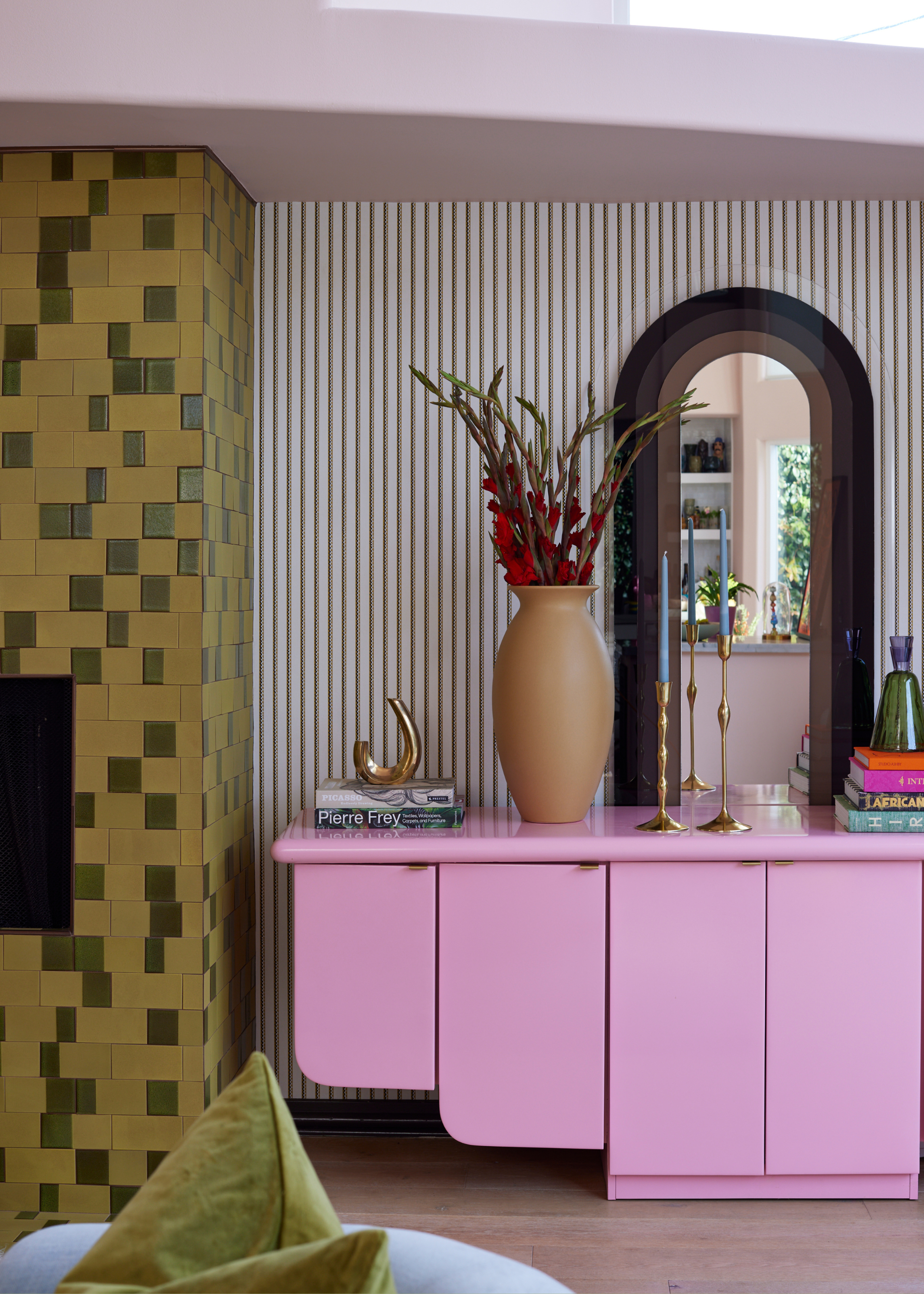 A living room with a mustard green and moss green tiled wall near a wall with striped wallpaper. A pink sideboard holding a vase of flowers, books and candle holders sits in front of an ovular mirror.