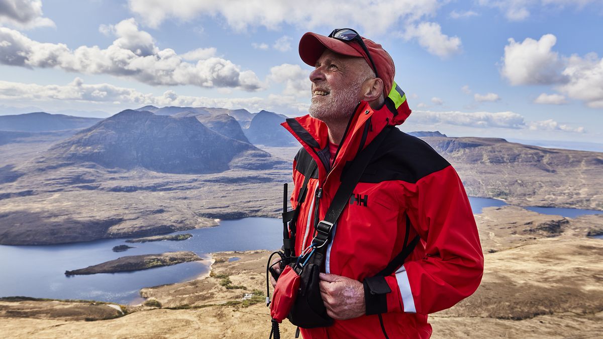 A mountain rescue volunteer in a bright red Helly Hansen jacket, against a background of mountains and lakes.