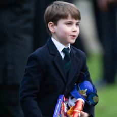 Prince Louis standing outside wearing a navy coat and blue and green plaid tie carrying a bunch of toys and smiling on Christmas 2024