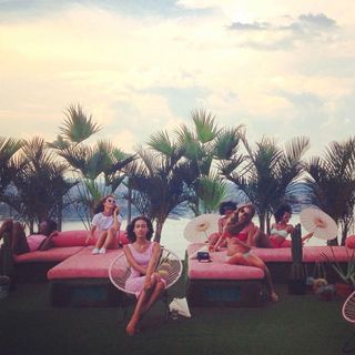 Models sitting on a pink lounge in front of the ocean and palm tree leaves