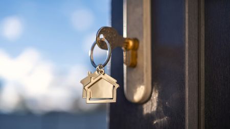 A close-up of a gold key and keychain shaped like a house. The key is in a home's lock. Conveys the ideas of renting, buying or selling a house, as well as mortgage rates.