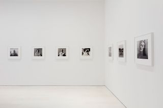 Portraits of different people in black & white photography are framed in white frames and hung on all white walls in the gallery.
