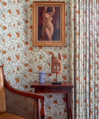 Close up of orange and green wallpaper and matching curtains, gold framed picture, dark wood side table and armchair