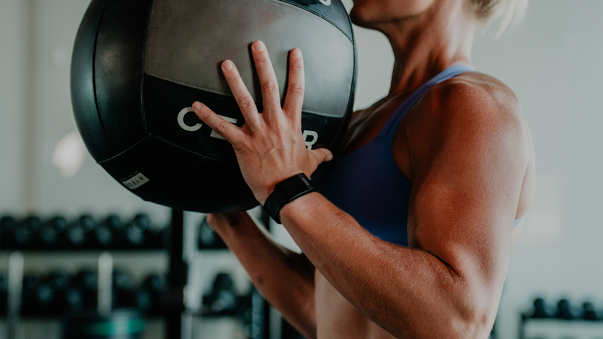 A close up of a woman lifting a large slam ball, a QALO SL-1 smart band on her wrist