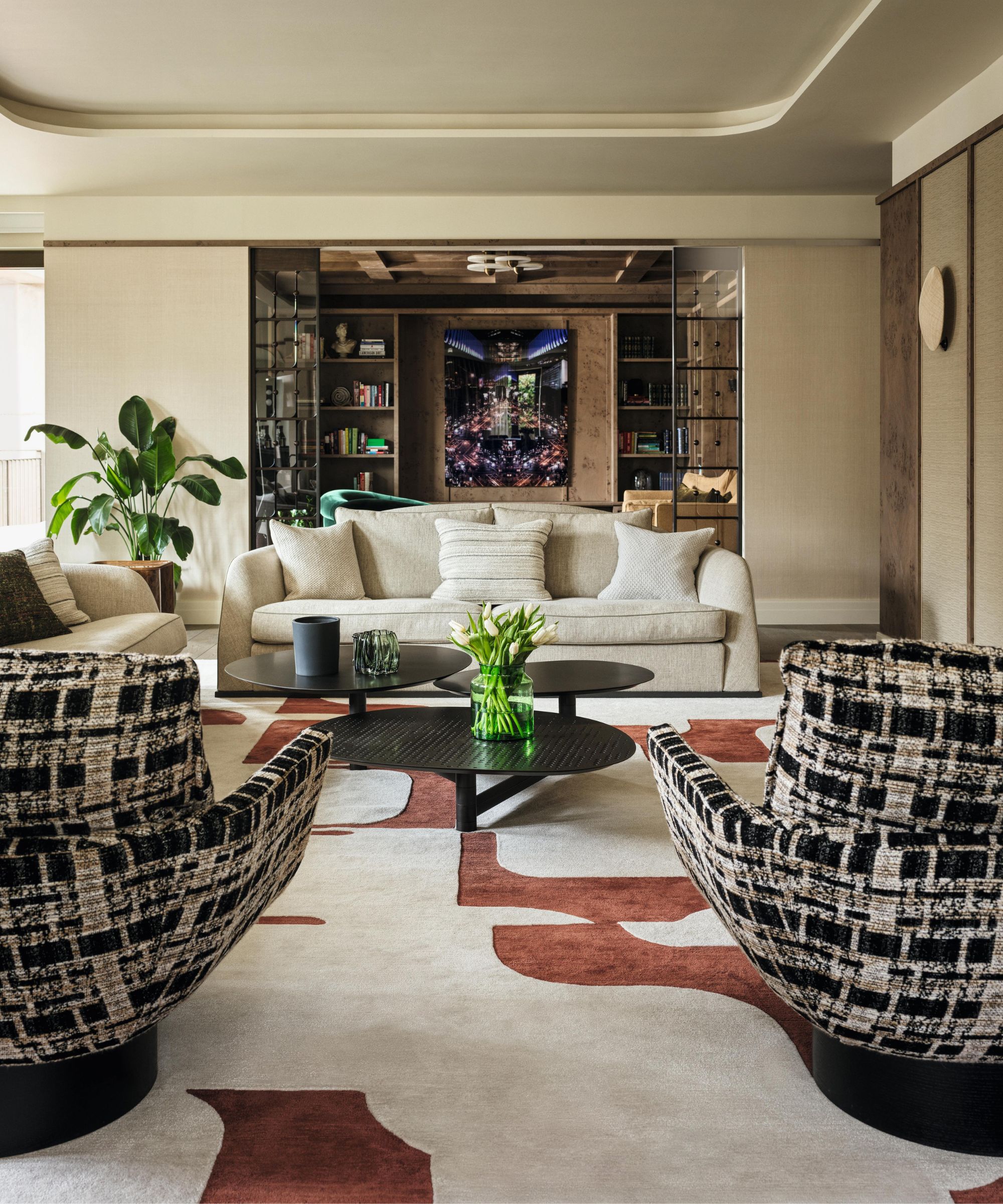a neutral sitting room with curved ceiling cornice detail, a large white and rust red patterned carpet, tweed armchairs and a neutral couch style with black round coffee tables and plants
