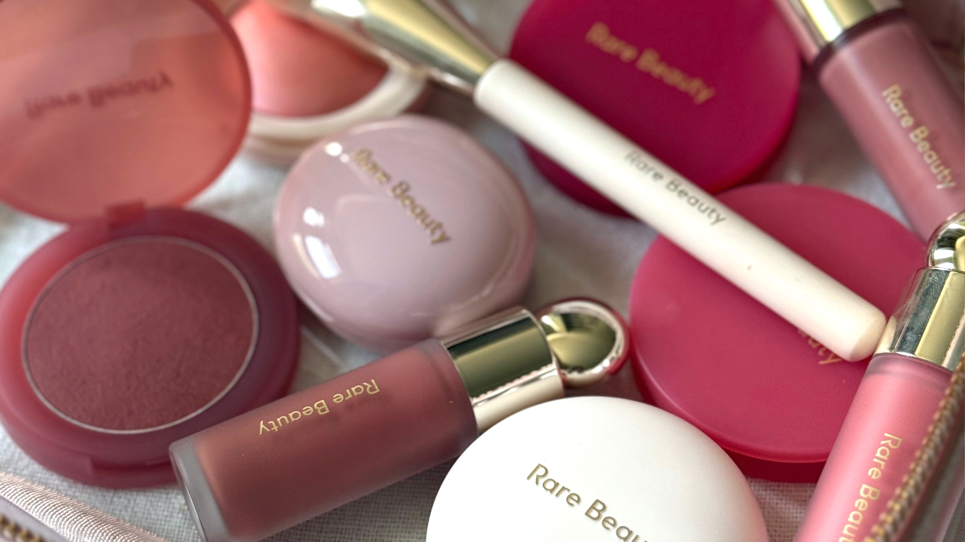 A close up shot of a selection of the various Rare Beauty blushers and a brush within a cosmetic bag