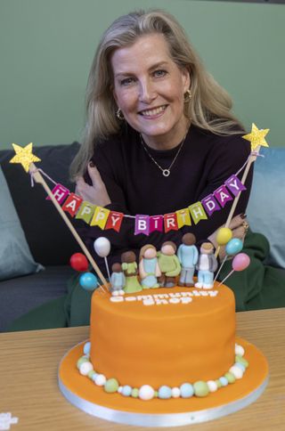 Duchess sophie wearing a maroon top and smiling in front of an orange cake that reads Happy Birthday with rainbow flags draped across it