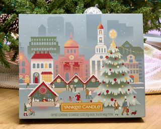 A Yankee Calendar advent calendar with muted pastel Christmas ice skating graphic against snowy city background, on floor under Christmas tree