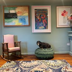 Dog sitting on patterned green pouffe by rocking chair and green wall with framed artworks