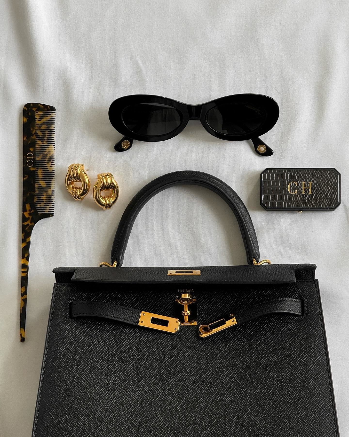 A flatlay picture of a black Hermes Kelly handbag, a tortoiseshell comb, gold earrings, black sunglasses and an embossed lip balm case with the initials CH