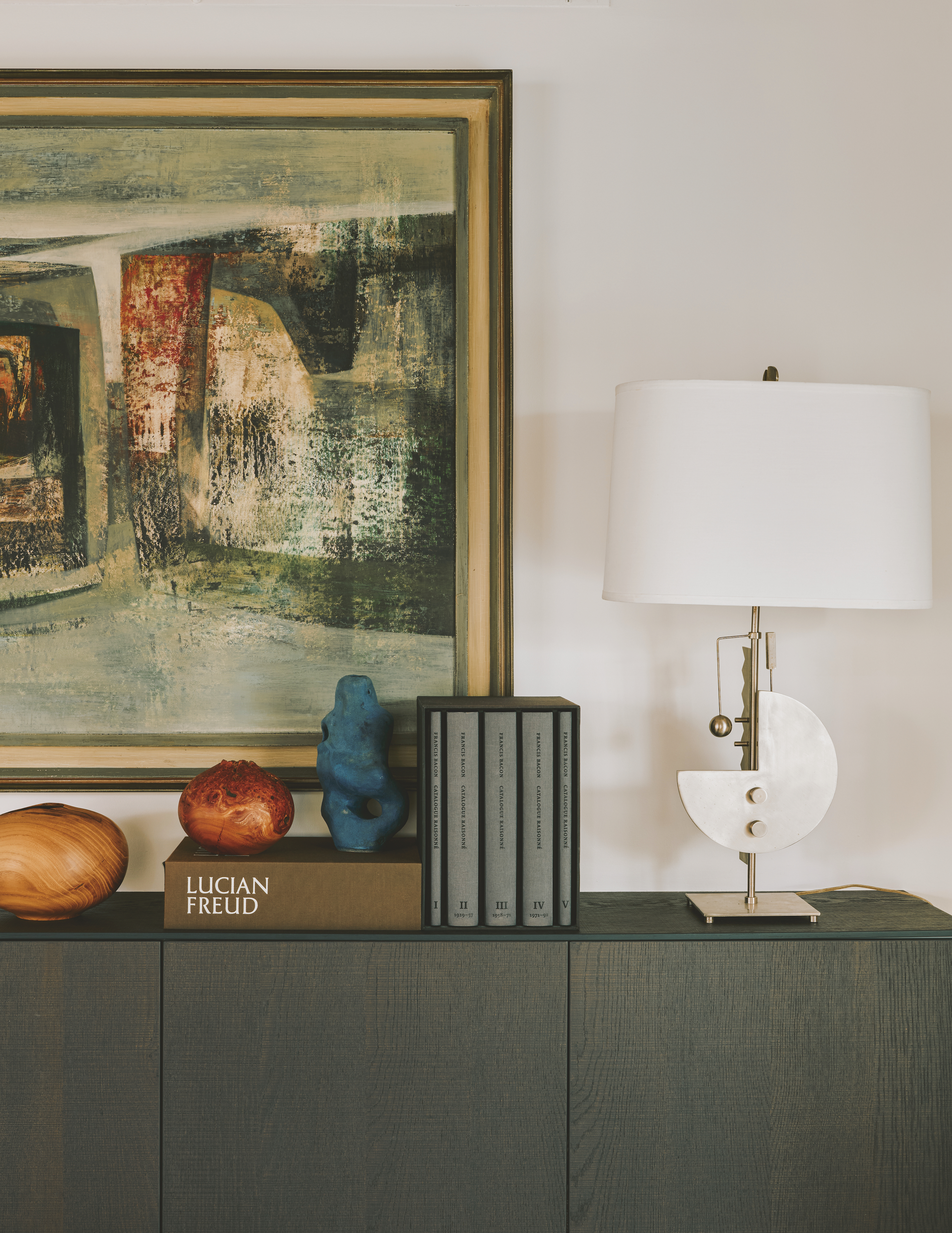 A dark wood side table with an artwork behind, some books and decor on top and a white, decorative lamp to the right.