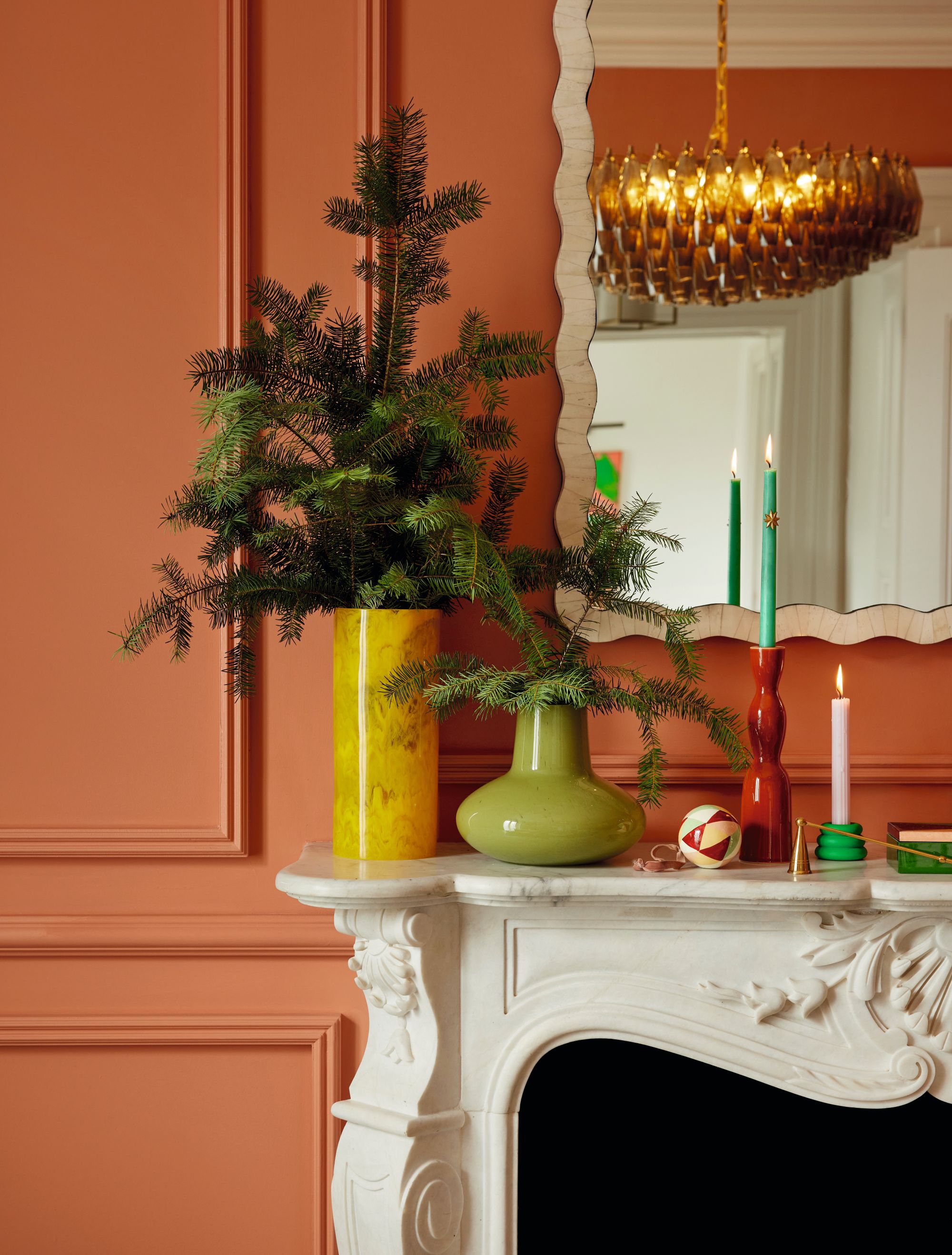 a white mantelpiece against a dark orange-painted paneled wall with a red and green vase on the mantelpiece filled with christmas tree branches next to lit candles in front of the wall hung mirror reflect an orange chandelier
