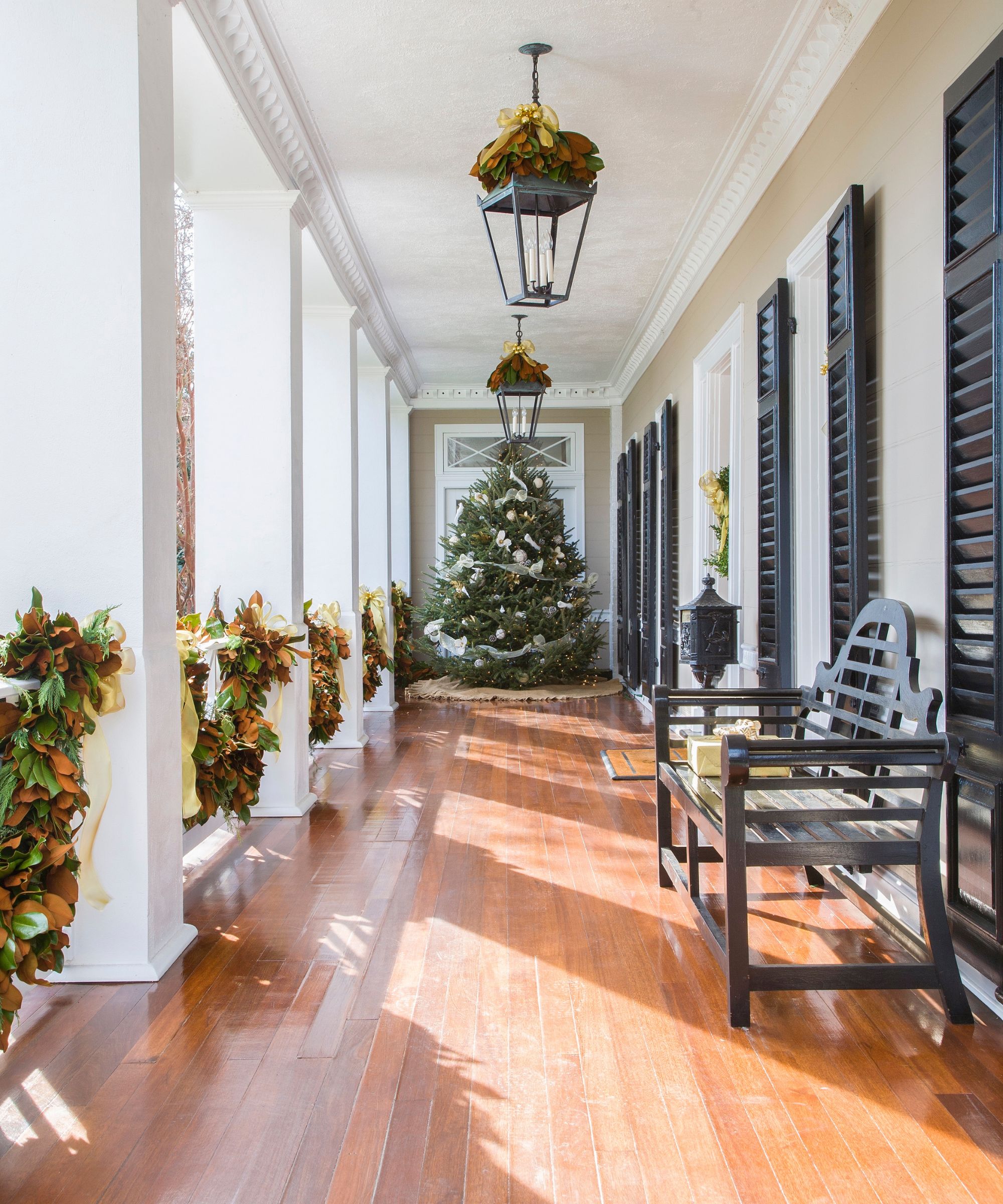 A long porch with white pillars and wooden flooring decorated for Christmas with a large tree, wreaths around the lights, and garlands draped over the banister