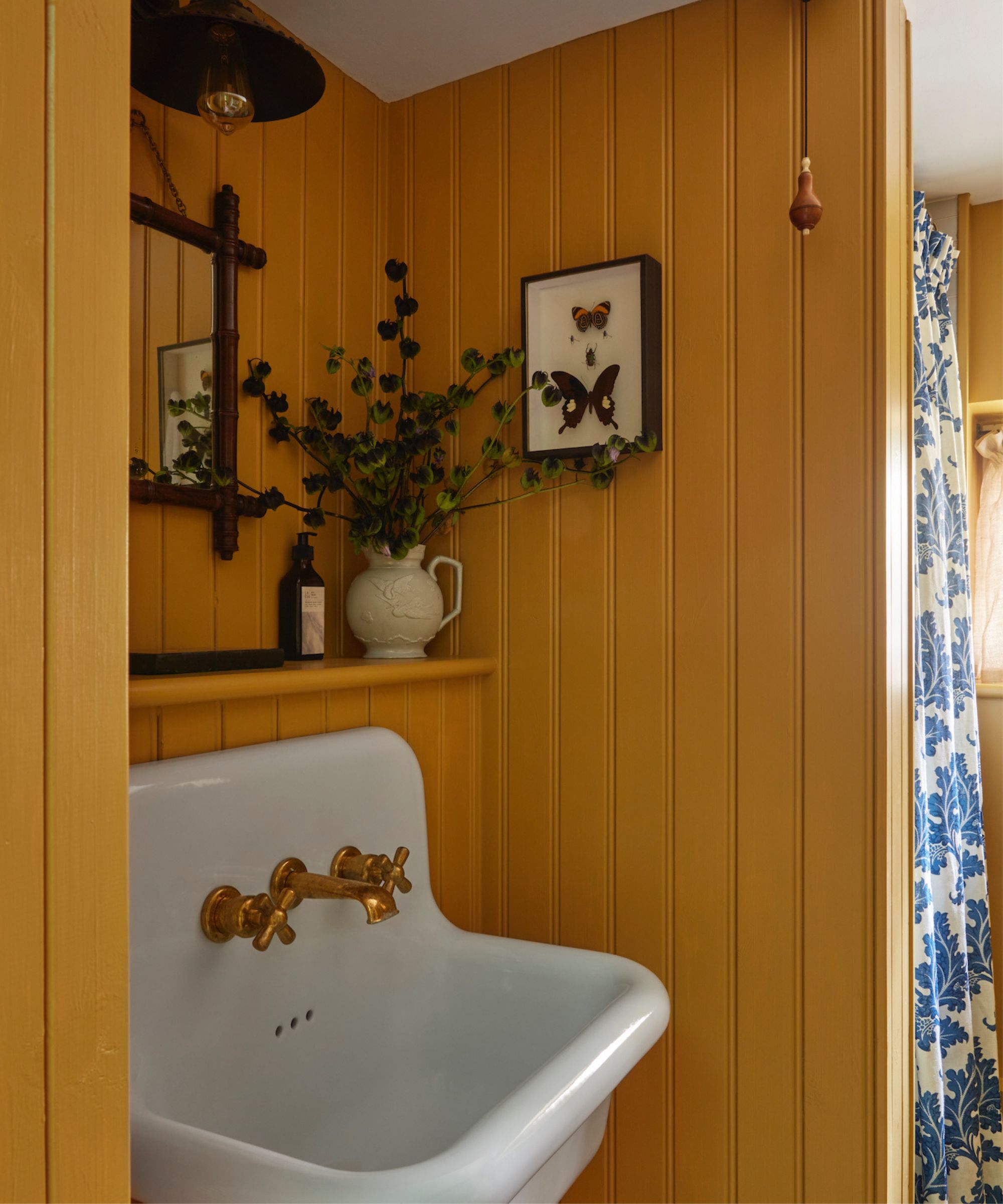 Powder room with panelled walls painted in Farrow &amp;amp; Ball's India Yellow, a ceramic sink with gold taps, and a vintage mirror with a pleated sconce above.