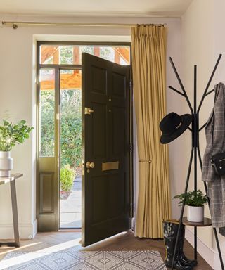 A hallway with an open dark green door, yellow curtains hanging to the right hand side of it, a table with a green plant on it to the left and a tall coat stand with hats on it to the right