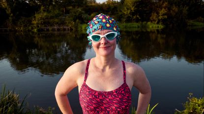 A woman in a swimsuit and colorful goggles stands in front of a lake.