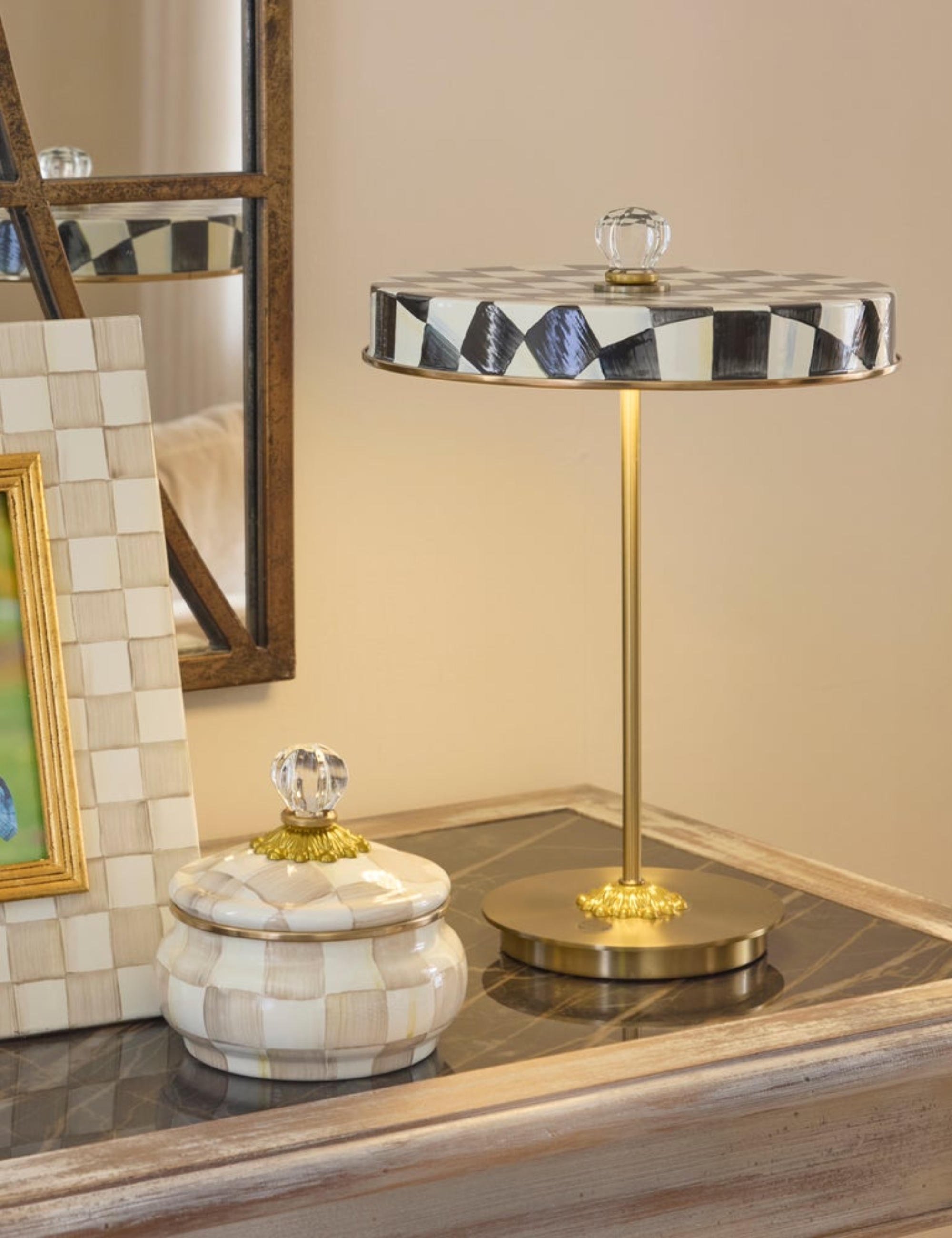 Mackenzie-Childs Courtly Check portable lamp in black-and-white styled alongside a frame and lidded vessel from the same collection in rose atop a marble credenza.