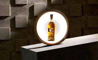 Bottle of whisky back lit in a circular framed display, grey stone square design wall, sloped stone slab display block