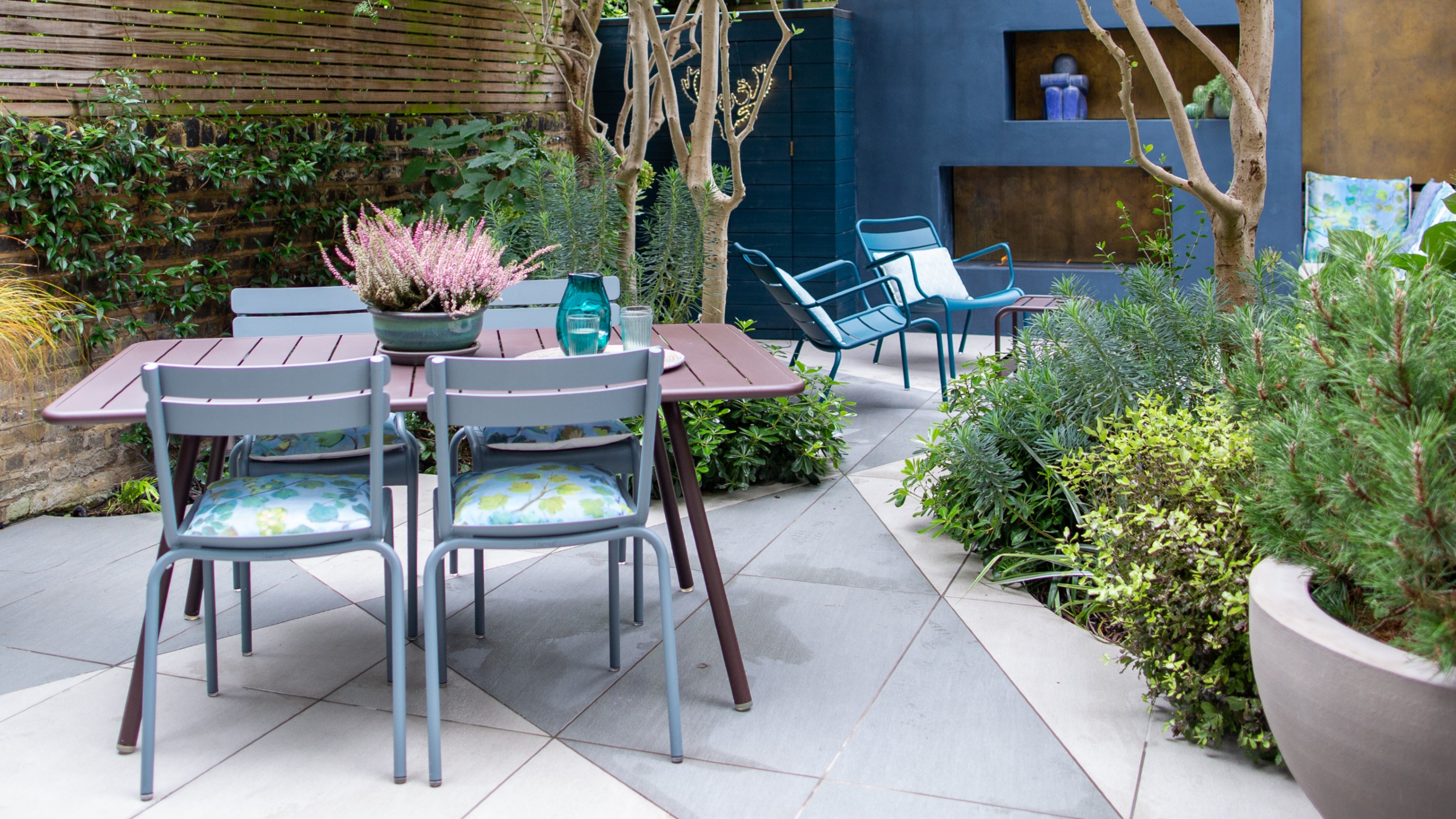 A paved garden with a dark red metal dining table and light blue dining chairs and two blue lounge chairs in the background