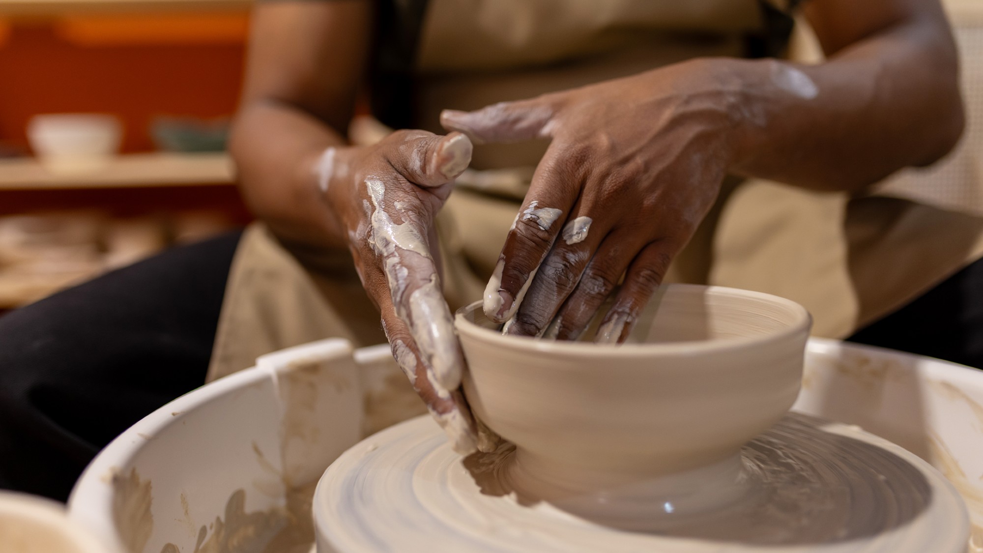 A craftsperson's clay-covered hands shape a wet clay bowl on a pottery wheel