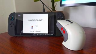 8Bitdo retro R8 gaming mouse next to Switch 2 running Mario Paint on woodgrain desk.
