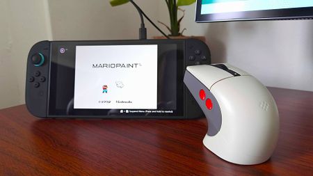 8Bitdo retro R8 gaming mouse next to Switch 2 running Mario Paint on woodgrain desk.