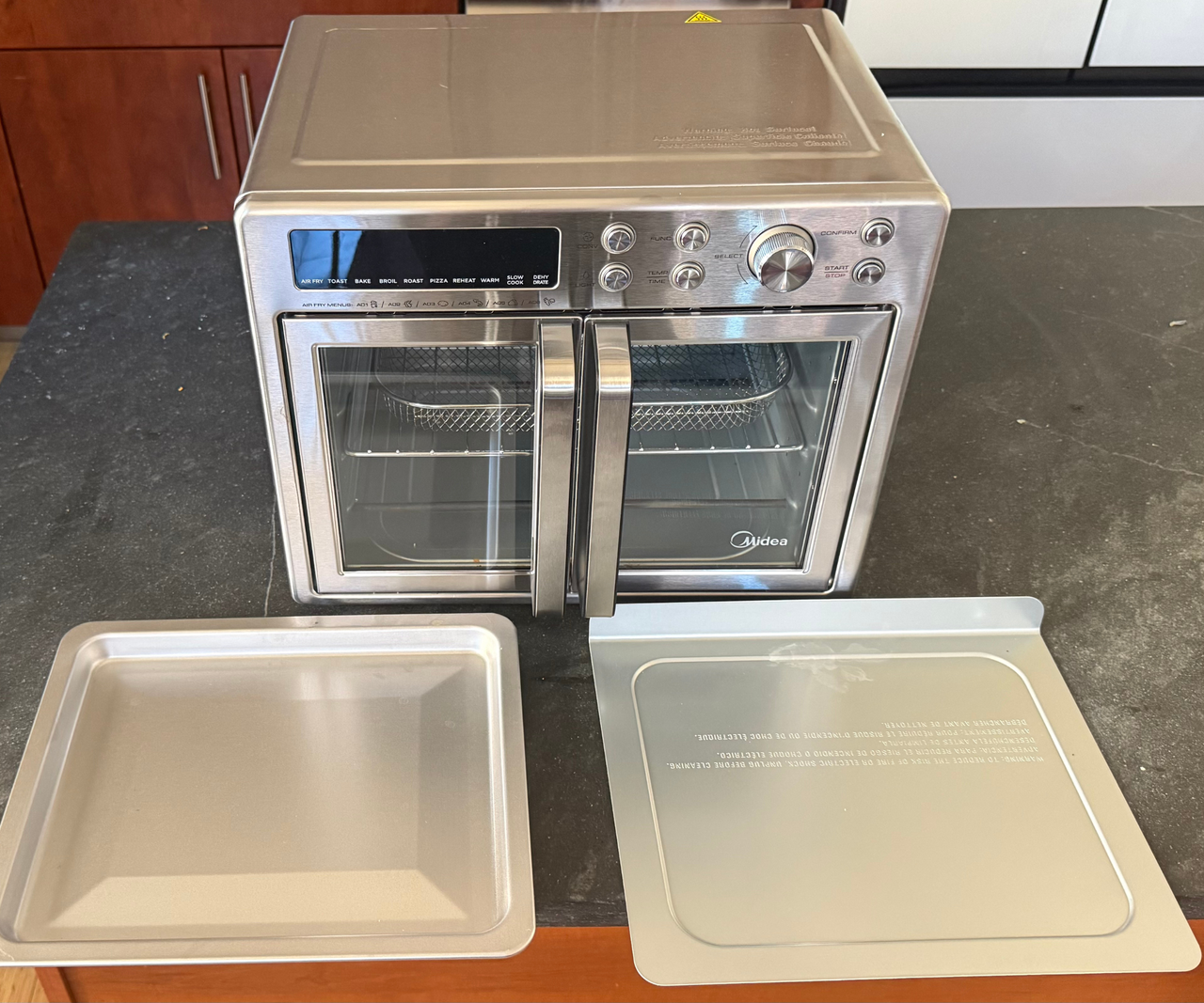 A stainless steel two door air fryer on a grey counter with two stainless steel baking trays in front of it.