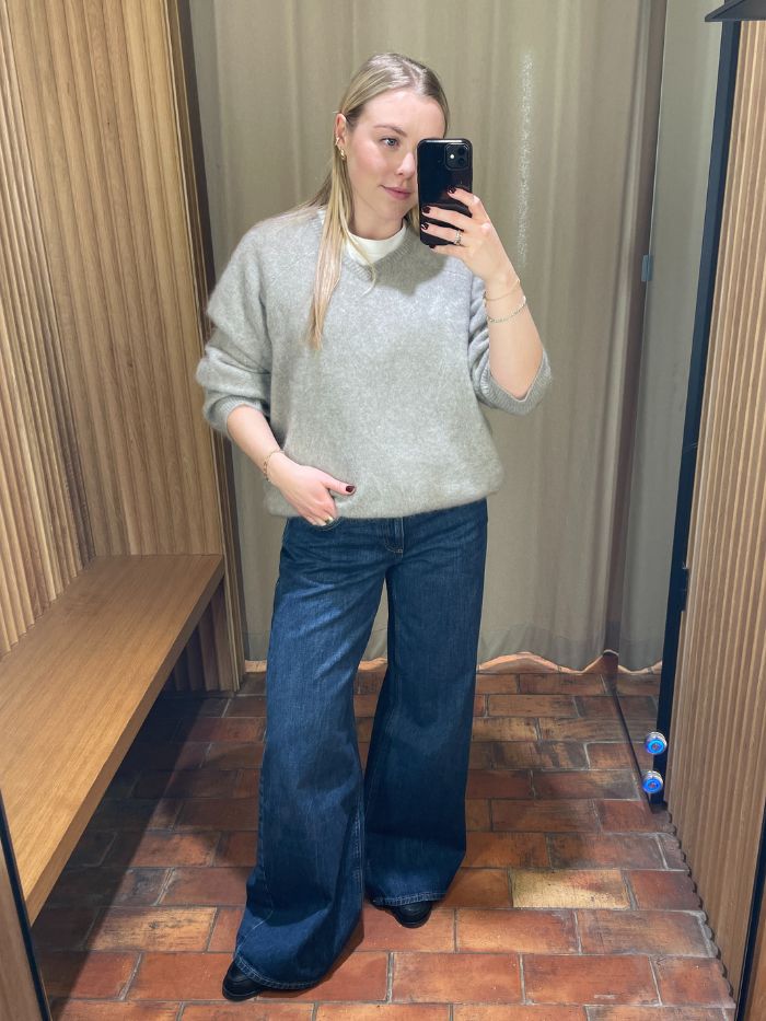 Woman wears grey jumper, blue wide leg jeans and loafers