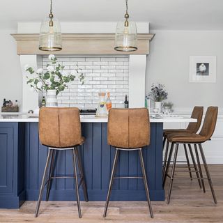 kitchen with wooden floorboards, blue island bench with white top, brown leather stools, white brick backsplash and hanging glass pendant lights