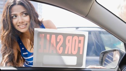 A woman tentatively looks into a car with a for sale sign in the driver's side window.