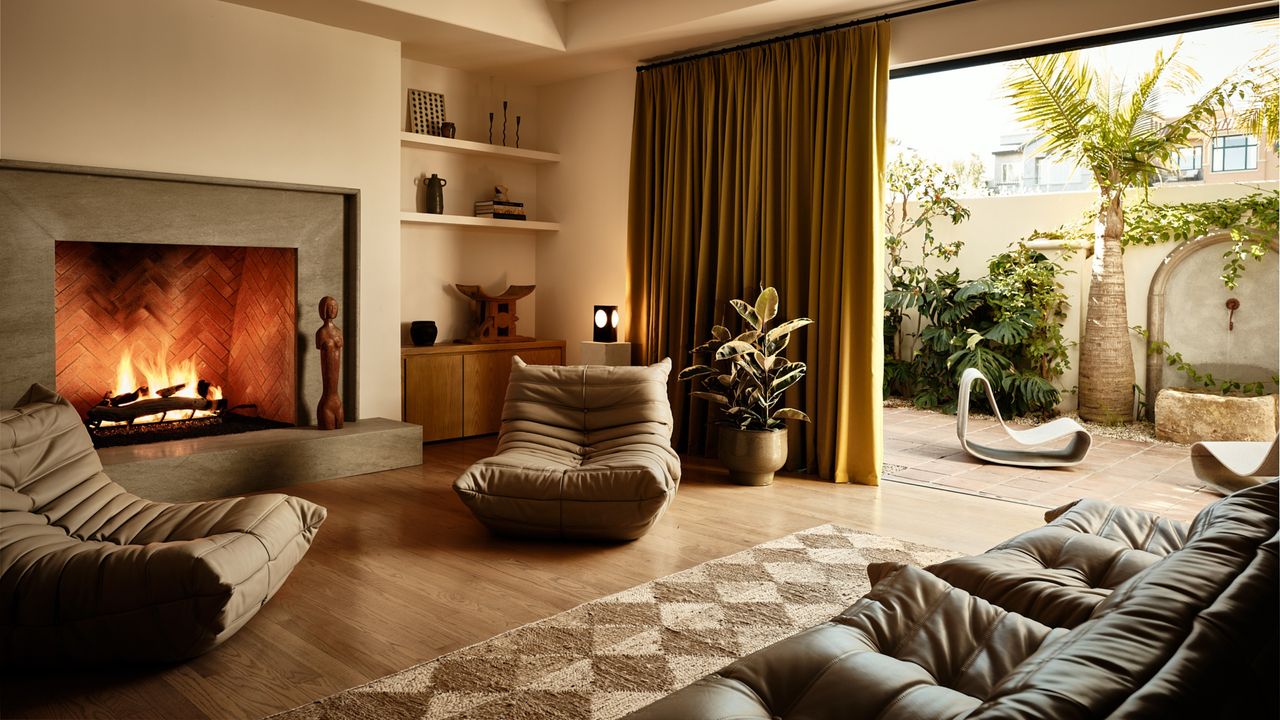 Image of a beige living room with a stone fireplace in the center of the main wall. There are two modern, brown accent chairs on either side of the fireplace and a checkerboard, jute rug. One wall is open to an outdoor space and there is an ochre yellow curtain pulled to the side.