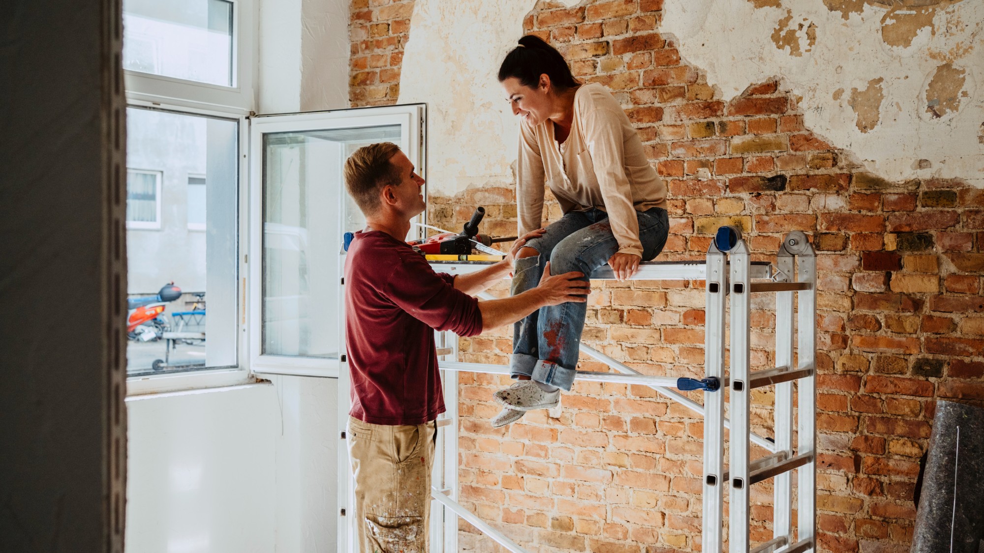 Man talking with woman sitting on a ladder in front of a brick wall during a home renovation