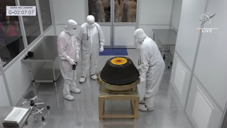 The capsule is seen in the clean room with scientists wearing white outfits gathered around.