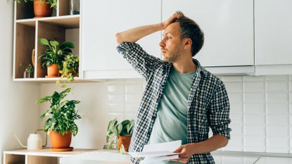 Worried man stands in kitchen holding tax papers