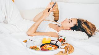 A woman lies on her back in bed with a tray of foods next to her that promote better sleep