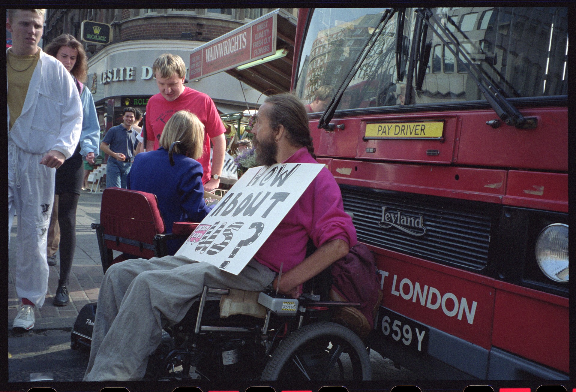 A group of disability rights protesters, several in wheelchairs, sit outside a London building holding handwritten signs, including one reading "Disabled people arrested trying to catch a bus! Why?"