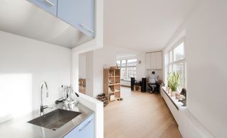 Open space in the flat, with white walls, ceiling and wooden floors. Image captured from the kitchen area with a view of a shoe shelf o nthe left, clear glass windows on the right (with pots of plants on the window sill