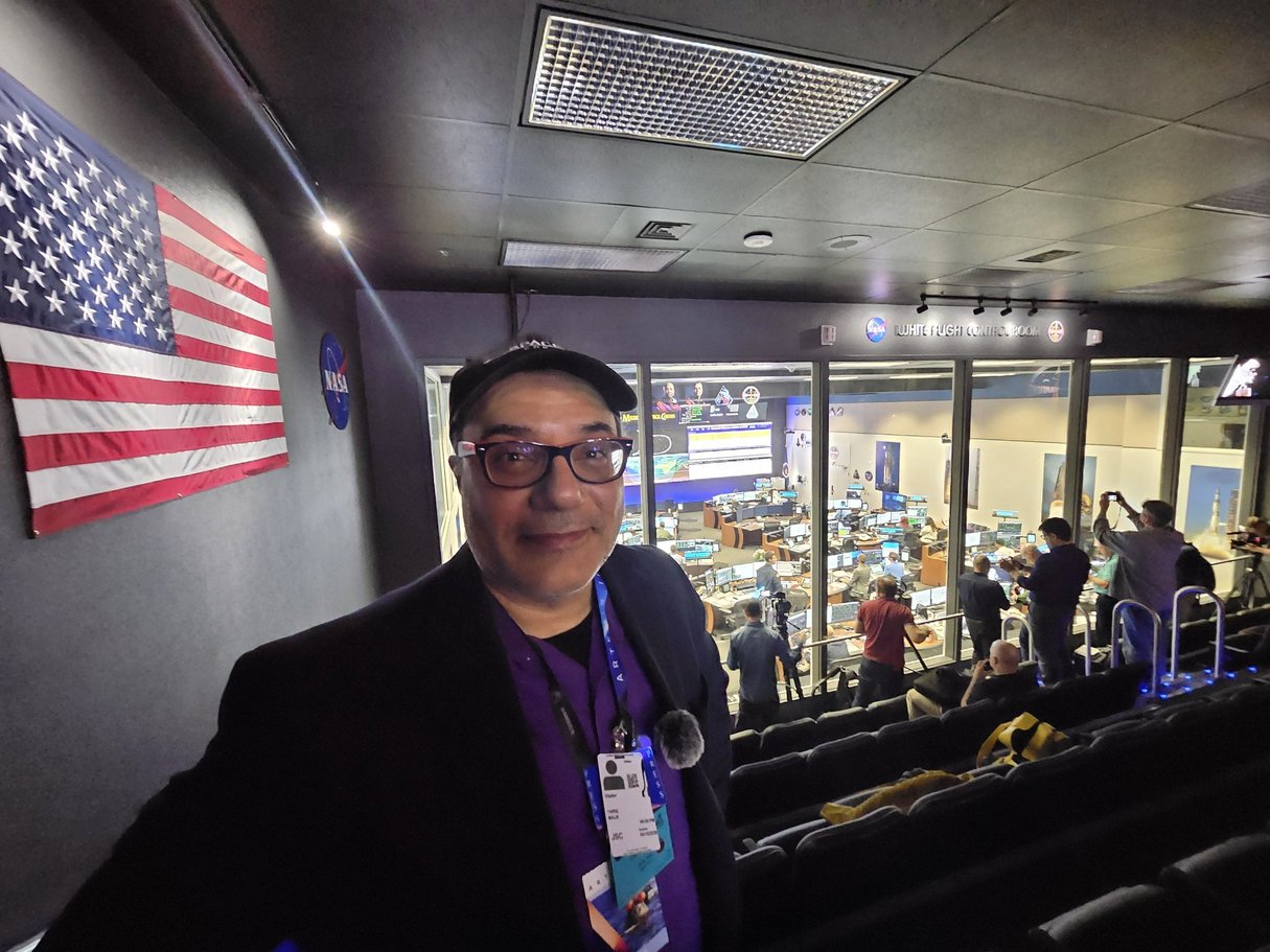 A man stands in a press room looking at the camera with an American flag on the wall to his left.