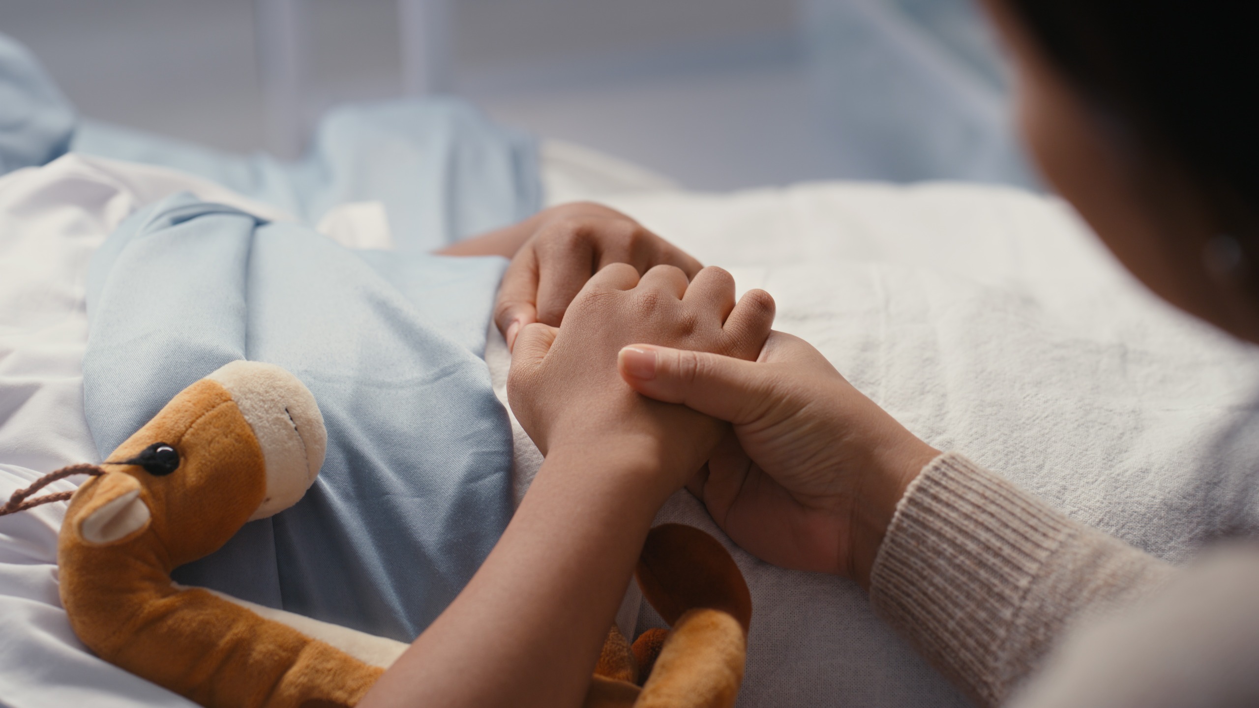close up on a child in a hospital bed holding a stuffed toy and their mother's hand