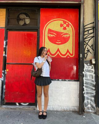 Aisha wears a short-sleeve white button-down, a pleated miniskirt, and black Mary-Jane flats while standing in front of a red-and-white wall